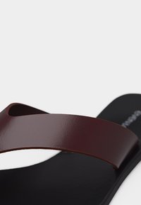 Καφέ δερμάτινο λουράκι πάνω σε μαύρη σόλα μοντέρνου σανδαλιού τύπου slip-on, το οποίο εμφανίζεται σε ανοιχτό γκρι φόντο.