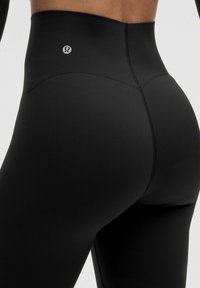 Højtaljede sorte leggings lavet af strækbart materiale med en glat finish, der har et lille logo ved taljen og flade sømme.