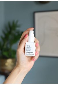 Biała butelka w sprayu z niebieskim napisem "spray na poduszkę głębokiego snu" od This Works, trzymana w ręce. Posiada dyszę pompkową i prostokątny kształt.