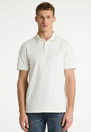 Jonge man met kort krullend haar draagt een wit poloshirt met zwarte biezen aan de kraag en mouwen, gecombineerd met een blauwe spijkerbroek.