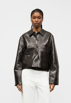 Woman wearing a cropped black leather button-up jacket with two front pockets and long sleeves, paired with white pants, standing against a plain background.