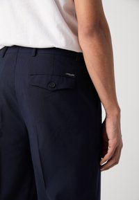 Mornarice s glatkom teksturom, s stražnjim džepom s kopčanjem na gumb i malom oznakom marke. Slim fit dizajn.