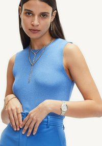 Gestreifter, ärmelloser Oberteil aus hellblauem Stoff, kombiniert mit geschichteter Schmuck in Goldtönen. Das Model trägt eine silberne Uhr, die das Outfit hervorhebt.