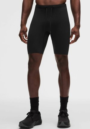 Mannelijk model draagt zwarte strakke sportshorts, zwarte crew-sokken en zwarte hardloopschoenen, staand tegen een effen achtergrond.