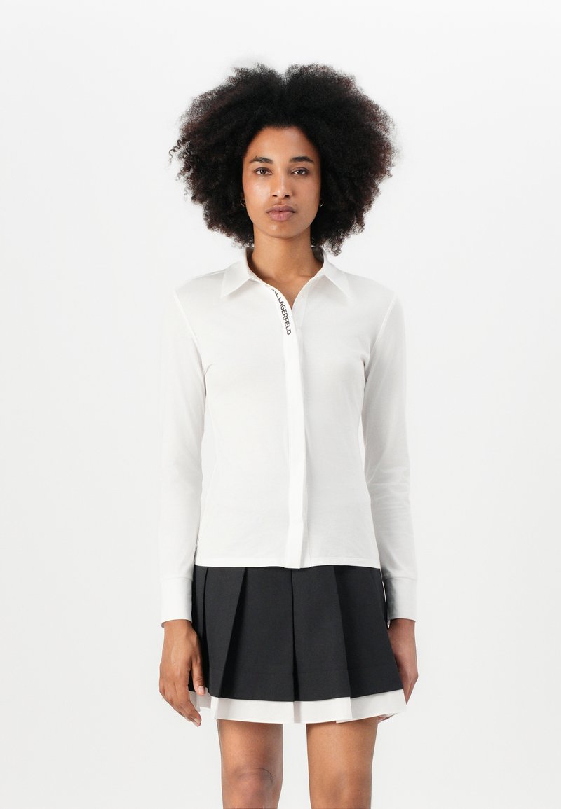 White long-sleeve shirt with a button-down collar, featuring a black logo along the front. Paired with a black pleated skirt.