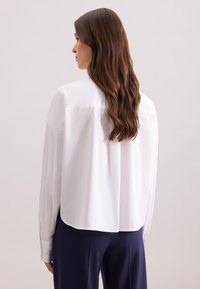 Camicia bianca a maniche lunghe con orlo arrotondato, dotata di spallina e polsini con bottoni. Abbinata a pantaloni blu navy, evidenziando una vestibilità comoda.