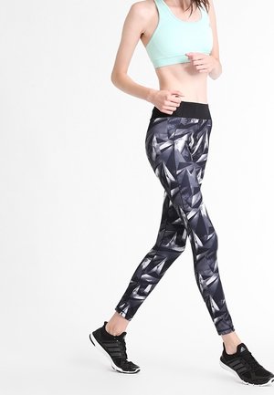 Leggings noir et gris à motifs avec une large taille noire, associés à un soutien-gorge de sport bleu clair et des chaussures de sport noires.