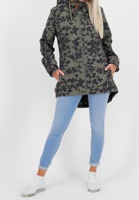 Vrouw in een groen en zwart bloemenjas met capuchon, lichtblauwe opgerolde skinny jeans, witte sneakers, met haar handen in de zakken van de jas.