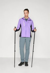 Vijolično-črna jakna z zadrgo in logotipom, sive hlače ter črni pohodni čevlji. Uporabnik drži treking palice, kar poudarja pohodniško opremo.