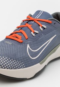 Scarpa da ginnastica blu e grigia con tomaia in rete testurizzata, lacci arancioni e logo Nike bianco. Presenta una suola ammortizzata.