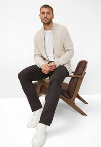 Beige gebreide cardigan met een rits, wit t-shirt, zwarte broek en witte sneakers. Zittend op een houten stoel met bruine accenten.