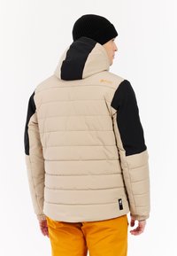 Donkerbeige pufferjack met zwarte mouwen, verstelbare capuchon, gestikte patronen en geribde boorden, gedragen met oranje broek. Logo op de achterkant weergegeven.