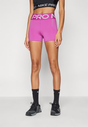 Femme portant un short Nike Pro taille haute violet vif, un soutien-gorge de sport noir Nike, des chaussettes noires Nike et des chaussures de sport noires, debout sur un fond blanc.