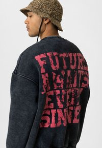 Donkergrijze oversized sweatshirt met in het groot roze tekst "TOEKOMST WACHT OP IEDEREEN SINDS" op de rug. Het model draagt een bucket hat met luipaardprint.