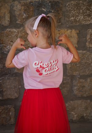 Junges Mädchen mit blonden Haaren im Pferdeschwanz, das ein pinkfarbenes "Cherry Club"-T-Shirt und einen roten Rock trägt und beide Arme vor einer Steinmauer anspannt.