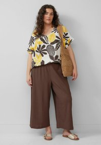 Bloes met bloemenpatroon in wit, geel en bruin. Bruine, wijde broek. Geweven bruine tas. Sandalen in lichte kleur met een accentgesp.