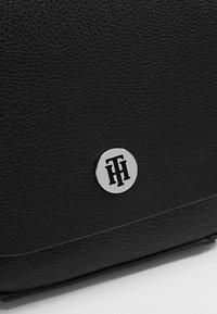 Black leather bag with textured finish, featuring a metallic circular logo accent on the front. Sleek and rectangular design.