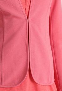 Zbliżenie różowej teksturowanej kurtki noszonej na jasnoróżowej bluzce, ukazujące otwarcie z przodu i zaokrąglone krawędzie.