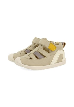 Calzado beige con correas de Velcro. Presenta laterales recortados, acentos en gris y amarillo en contraste, y suelas de goma para mayor agarre.