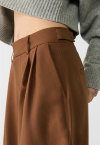 Hnědé kalhoty na míru s sklady v přední části, které mají poutka na opasek a boční kapsu, vystavené na světlém pozadí.