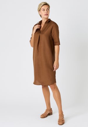 Mujer rubia de pie con un vestido marrón hasta la mitad de la pierna con mangas cortas y sandalias trenzadas de color beige, mirando hacia su izquierda sobre un fondo blanco.