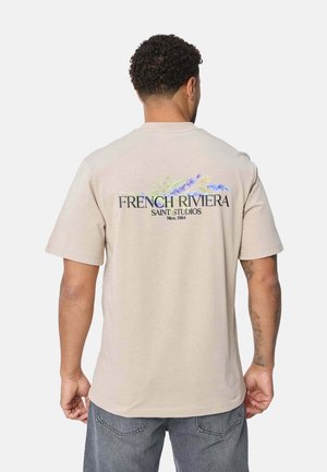 T-shirt en coton beige à manches courtes, avec le texte "FRENCH RIVIERA" et des accents floraux au dos. Coupe standard.