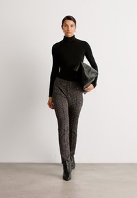 Un haut noir à col roulé associé à un pantalon slim noir et blanc à motifs, tenant un sac à main noir texturé. Des bottines noires à la cheville complètent le look.