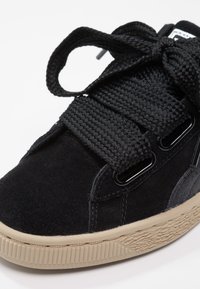 Gros plan d'une basket en daim noir avec des lacets tissés noirs épais et une semelle en caoutchouc beige texturée.