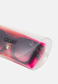 Gafas de sol con lente de degradado de púrpura a negro en un estuche de plástico transparente con un interior rosa y cierre a presión dorado. Nombre de la marca "QUAY" visible.