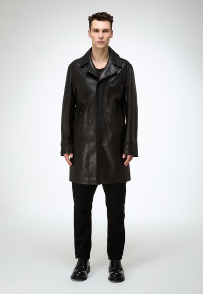 Schwarzer Ledermantel mit Reverskragen, zwei Seitentaschen und geradem Schnitt. Wird über schwarzen Hosen und Schuhen getragen.