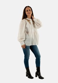 Blusa bianca a maniche lunghe con ricami dorati e dettagli plissettati, abbinata a jeans aderenti blu e stivali neri con tacco alto.