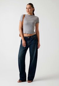 Grijs cropped t-shirt gecombineerd met losse, donkerblauwe spijkerbroek. Model draagt bruine sandalen en heeft een bruine schoudertas bij zich.