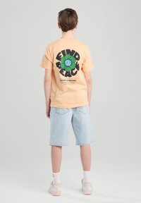 Persikka lyhythihainen t-paita, jossa on suuri musta graafinen painatus vihreällä ja valkoisella tekstillä takana, yhdistettynä vaaleansinisiin denim-shortsihousuihin.