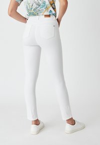 Jeans blancos ajustados con cintura alta, que cuentan con dos bolsillos traseros y una etiqueta de piel marrón en la cinturilla, combinados con zapatillas blancas.