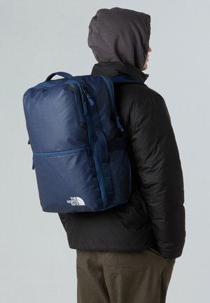 Sac à dos bleu marine avec surface texturée, double fermeture éclair et bretelles ajustables. Présente un logo sur le bas à l'avant et une forme rectangulaire.