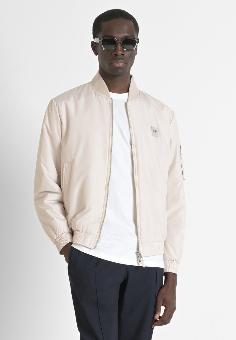 Chaqueta bomber beige con un acabado suave, cierre de cremallera y puños elásticos. Camiseta blanca debajo. Modelo con gafas de sol oscuras.