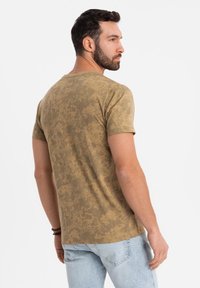 Khaki tričko s texturovaným, skvrnitým vzorem; krátké rukávy a design s kulatým výstřihem. Nosené s světle modrými džíny.