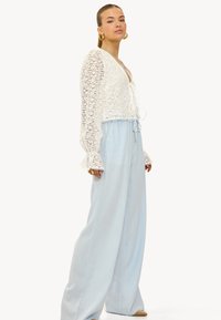 Haut en dentelle blanche à manches longues évasées, associé à un pantalon bleu clair ample en tissu doux et texturé. Les détails incluent une taille avec cordon de serrage.