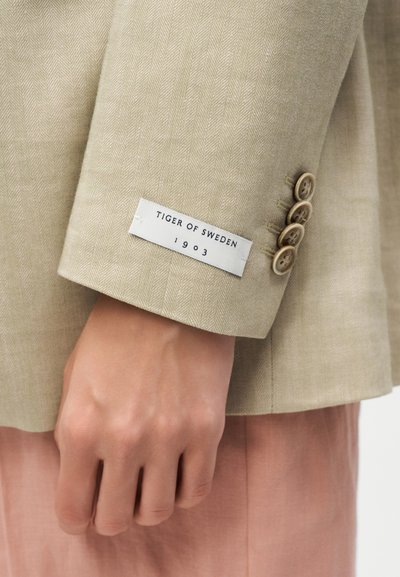Μαύρη μανσέτα μπεζ blazer με τέσσερα κουμπιά και ετικέτα "Tiger of Sweden 1903" πάνω από ένα χέρι που ακουμπά κοντά σε ροζ παντελόνια.