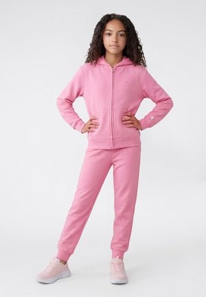 Fille aux cheveux bouclés, debout les mains sur les hanches, portant un sweat à capuche rose avec fermeture éclair, un pantalon de jogging assorti et des baskets rose clair.