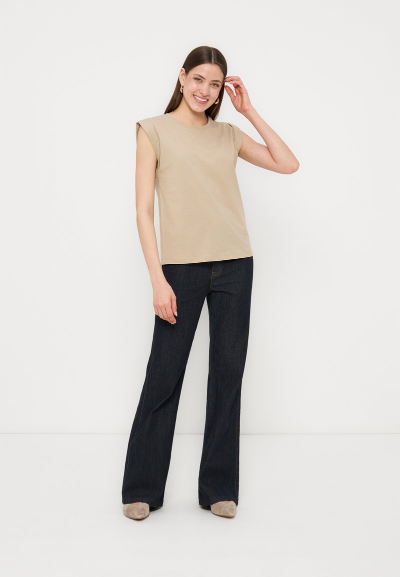 Χαμογελαστή γυναίκα με μακριά σκούρα μαλλιά φοράει μπεζ αμάνικη μπλούζα με βάτες στους ώμους, σκούρο παντελόνι με φαρδιά πόδια, μπεζ παπούτσια, στέκεται σε λευκό φόντο.