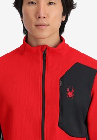 Giacca rossa con zip realizzata in tessuto testurizzato, presenta un pannello nero con una tasca con zip e un logo di ragno rosso sul petto.