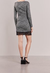 Robe grise en chevrons à manches longues avec une silhouette ajustée, détail de bord en dentelle, associée à des chaussures à talons carrés en cuir verni noir.