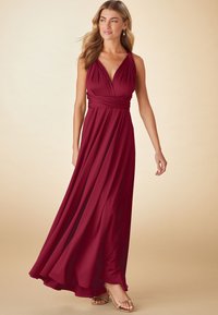 Kvinde iført en ærmeløs, dybrød, gulvlængde kjole med indsnøret talje, stående med et blødt smil mod en beige baggrund.