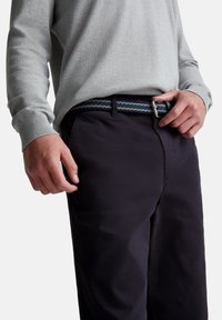 Hombre ajustando un cinturón tejido en azul, beige y marino, llevado con pantalones oscuros y un suéter de manga larga gris claro, frente a un fondo blanco.