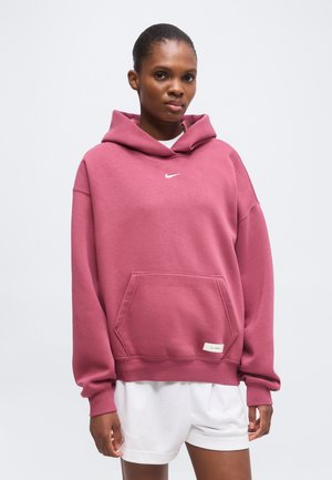 Personne portant un sweat à capuche rose ample avec un logo Nike blanc et un short en mesh blanc, debout devant un fond clair uni.