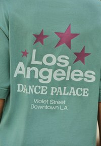 Tricou din bumbac verde mentă, cu text mare alb: "Palatul Dansului Los Angeles" și stele de culoare burgund. Include detalii despre locație dedesubt.