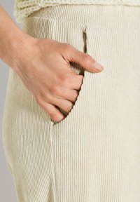 Hand in de zak van crème-kleurige ribcord broek, met een deel van een gebreide crème trui zichtbaar.