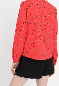 Czerwona bluzka z długim rękawem z czarno-niebieskim okrągłym wzorem; ma zgromadzone ramiona i zaokrąglony dół. W połączeniu z czarnymi szortami.