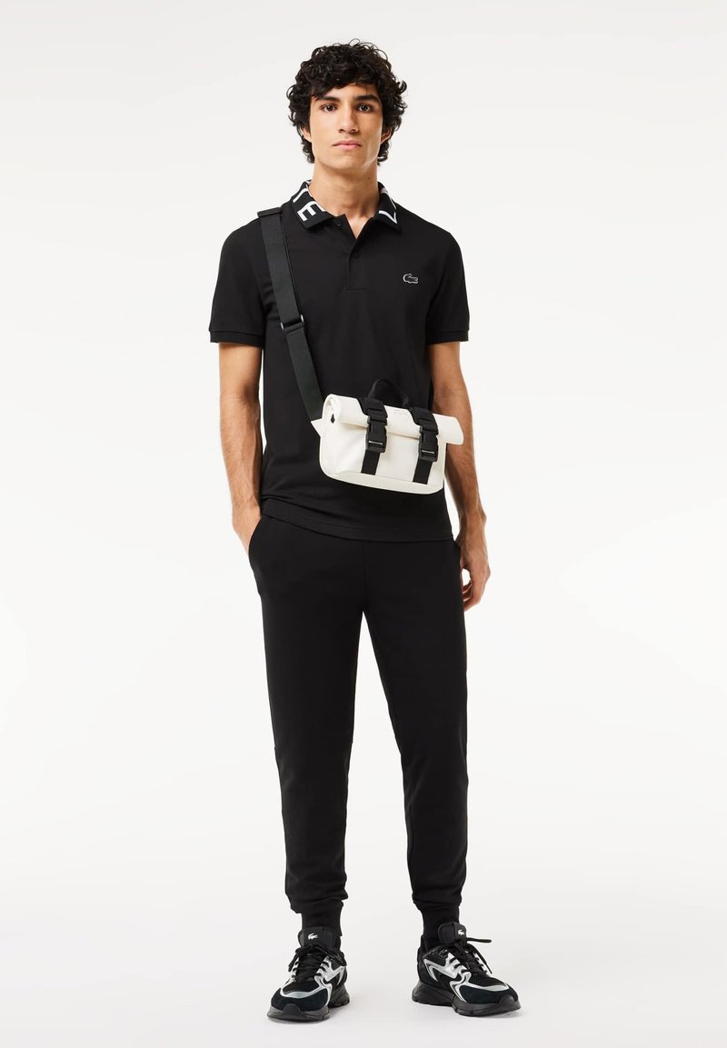 Schwarzes Polo mit weißen Akzenten, schwarze Jogginghose und schwarze Sneakers. Eine weiße Umhängetasche mit schwarzen Riemen und Schnallen rundet das Outfit ab.